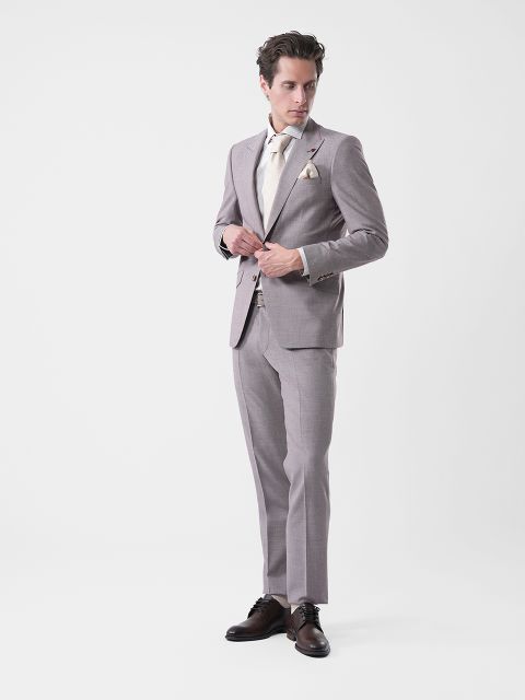 Mann in hellgrauem Anzug, cremefarbenem Hemd und Krawatte, der vor einem schlichten weißen Hintergrund steht, seine Jacke zuknöpft und zur Seite schaut.