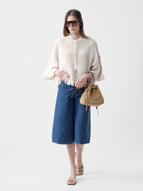 Frau trägt eine Sonnenbrille, einen cremefarbenen Strickpullover mit Fransen, blaue knielange Jeans-Culottes und Sandalen und hält eine hellbraune Handtasche vor einem weißen Hintergrund.