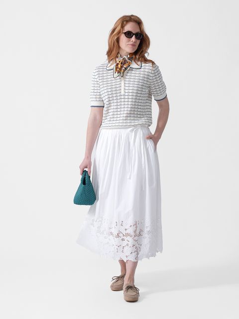 Frau trägt eine Sonnenbrille, ein gestreiftes Oberteil, einen weißen Midirock mit Stickerei, einen Schal um den Hals, eine blaugrüne Tasche und beige Schuhe.