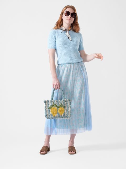 Frau mit Sonnenbrille, die ein hellblaues Oberteil, einen blauen Rock mit Blumenmuster, braune Sandalen und einen Schal trägt und eine gewebte Tasche mit gelben Zitronen in der Hand hält.