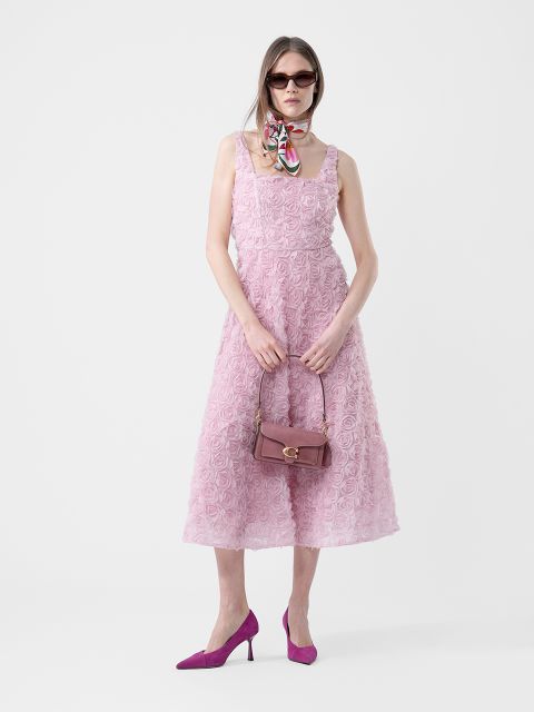 Frau in einem ärmellosen rosa strukturierten Kleid, lila Absätzen, Sonnenbrille, einem Schal um den Hals und mit einer lila Handtasche in der Hand, stehend vor einem weißen Hintergrund.