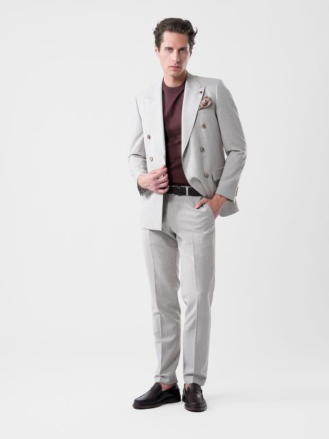 Ein Mann in einem hellgrauen Anzug und kastanienbraunem Hemd posiert selbstbewusst vor einem weißen Hintergrund, eine Hand an der Jacke, die andere in der Tasche.