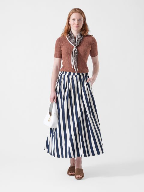 Frau mit braunem Oberteil, gestreiftem Maxirock, Schal und Sandalen, die eine kleine weiße Handtasche in der Hand hält, vor weißem Hintergrund.