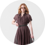 Frau mit Sonnenbrille und einem kurzärmeligen, gegürteten braunen Kleid, die mit einer Hand in der Hüfte vor einem hellen, runden Hintergrund steht.
