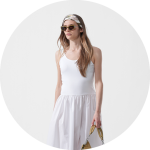 Frau mit Sonnenbrille und weißem Kleid mit weißem Stirnband, die eine hellbraune Handtasche in der Hand hält, steht vor einem schlichten hellen Hintergrund.