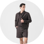 Ein Mann in dunkelbrauner Jacke, Polohemd und Shorts steht vor einem hellgrauen Hintergrund und blickt mit einer Hand auf seiner Jacke zur Seite.