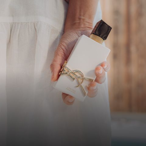 Eine Person in einem weißen Kleid hält eine weiße Parfümflasche mit goldenen Akzenten und einer schwarzen Kappe vor einem unscharfen braunen Hintergrund.