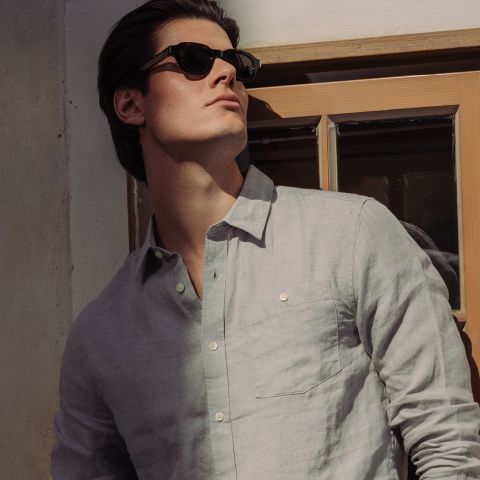 Ein Mann mit Sonnenbrille und hellgrauem Button-up-Hemd steht am Fenster und blickt in die Sonne.