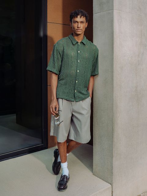 Ein Mann in einem grünen kurzärmeligen Hemd, grauen Shorts, schwarzen Halbschuhen und weißen Socken steht an einer Betonmauer und trägt eine Sonnenbrille.