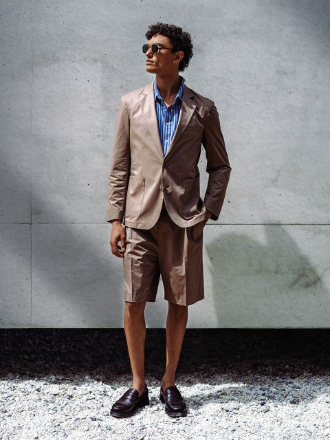 Ein Mann, der eine Sonnenbrille, einen hellbraunen Blazer mit passenden Shorts, ein blau gestreiftes Hemd und Halbschuhe trägt, steht an einer Betonmauer auf Kies.