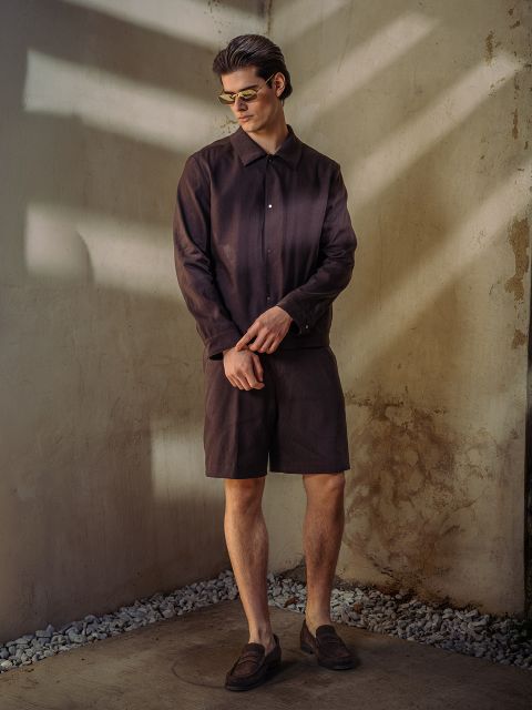 Ein Mann mit Sonnenbrille, dunkelbraunem Button-up-Hemd, passenden Shorts und Halbschuhen steht vor einer sonnenbeschienenen, strukturierten Wand mit Steinakzenten.
