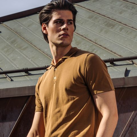 Ein Mann in einem braunen, kurzärmeligen Poloshirt steht draußen vor einem Metalldach und schaut mit ernster Miene zur Seite.