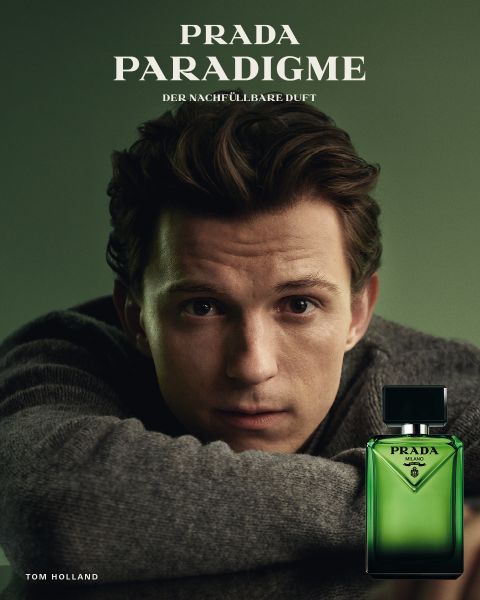 Ein Mann stützt sein Kinn auf die Hand neben einem grünen Prada Paradigme-Parfümflakon mit dem Produktnamen und dem Text "Tom Holland" auf dem Bild.