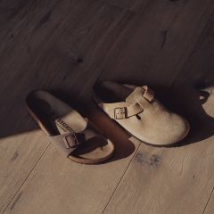 Zwei nicht zusammenpassende Birkenstock-Sandalen - eine mit offenen und eine mit geschlossenen Zehen - mit Schnallen auf einem sonnenbeschienenen Holzboden.
