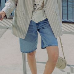 Eine Person, die einen hell gestreiften Blazer, eine bedruckte Bluse und Jeansshorts trägt, steht im Freien an einem Geländer und hält eine kleine beigefarbene Handtasche in der Hand.