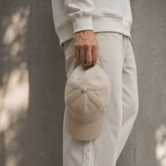 Eine Person, die helle Kleidung trägt, hält eine beigefarbene Baseballmütze vor einem neutralen, strukturierten Hintergrund an ihrer Seite.