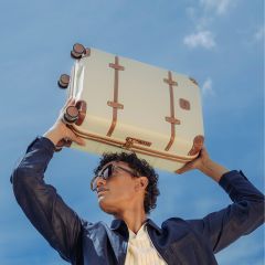 Eine Person mit Sonnenbrille und dunkler Jacke hält einen cremefarbenen Koffer mit braunen Riemen über ihren Kopf vor einem blauen Himmel.