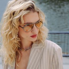 Eine Frau mit lockigem blondem Haar, die eine getönte Flieger-Sonnenbrille, einen gestreiften beigen Blazer und eine klobige Kettenhalskette trägt, steht an einem Gewässer.