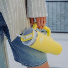 Person in Jeansshorts und gestreifter Jacke, die einen großen gelben Stanley-Becher mit Strohhalm und Henkel hält.