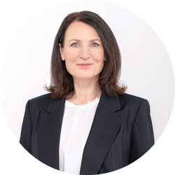 Frau mit schulterlangen braunen Haaren, die einen schwarzen Blazer und eine weiße Bluse trägt, lächelt sanft vor einem schlichten hellen Hintergrund.