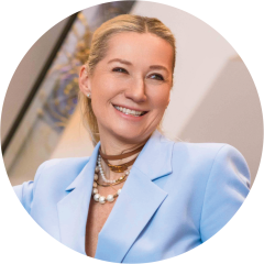 Lächelnde Frau mit blondem Haar in einem Pferdeschwanz, die einen hellblauen Blazer, mehrlagige Halsketten und Perlenohrringe trägt.