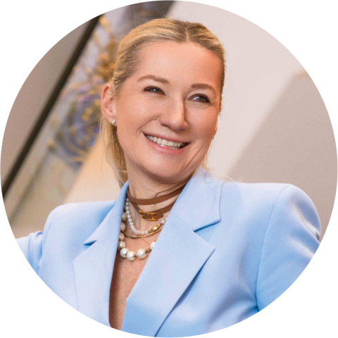 Lächelnde Frau mit blondem Haar in einem Pferdeschwanz, die einen hellblauen Blazer, mehrlagige Halsketten und Perlenohrringe trägt.