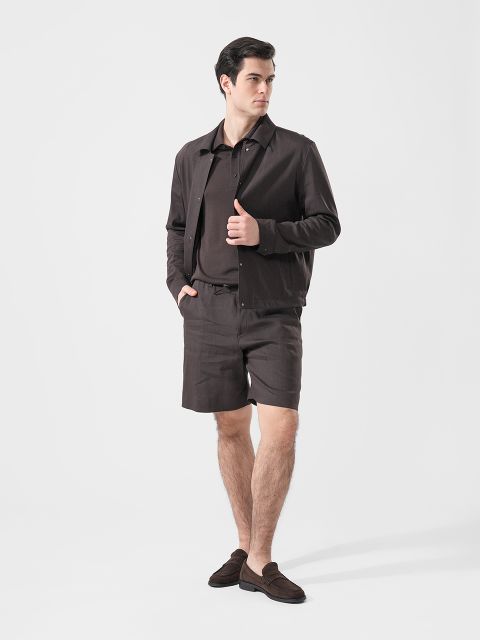 Mann in brauner Jacke, passendem Polohemd, Shorts und Halbschuhen vor einem schlichten weißen Hintergrund.
