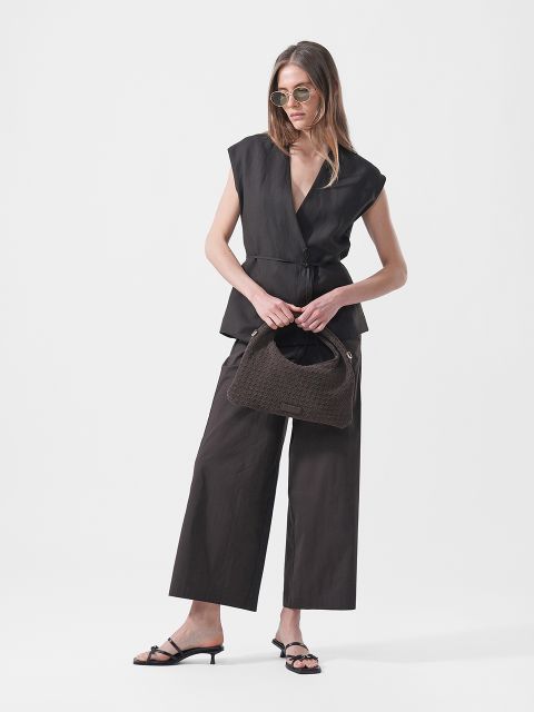 Frau trägt eine Sonnenbrille, ein ärmelloses schwarzes Wickeloberteil, eine schwarze Hose mit weitem Bein und hochhackige Sandalen und hält eine braune Handtasche mit Struktur vor einem weißen Hintergrund.