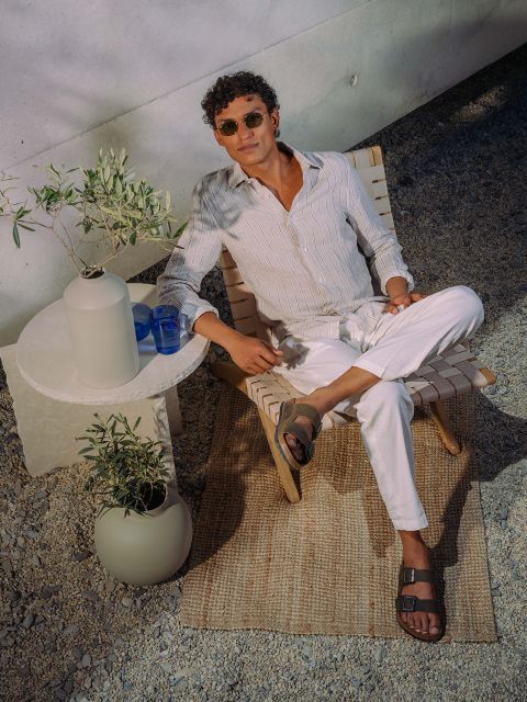 Ein Mann mit Sonnenbrille und leichter Kleidung entspannt sich auf einem Liegestuhl neben einem kleinen Tisch mit Pflanzen und Gläsern, draußen auf einem Kieselsteinboden und einem gewebten Teppich.