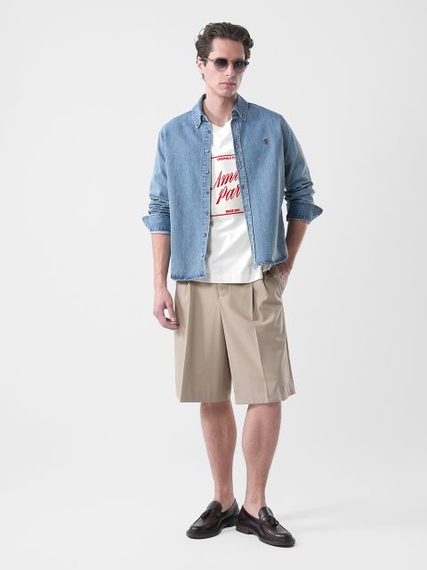 Mann, der eine Sonnenbrille, ein Jeanshemd über einem grafischen T-Shirt, beigefarbene knielange Shorts und braune Halbschuhe trägt und vor einem schlichten Hintergrund steht.