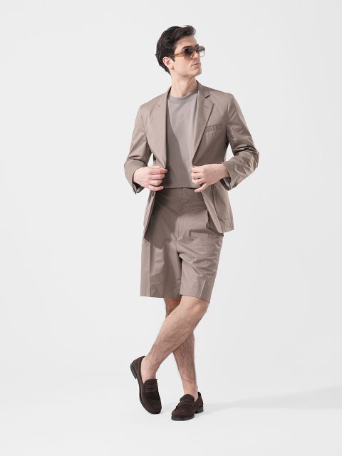 Ein Mann, der eine Sonnenbrille, einen taupefarbenen Blazer, passende Shorts, ein graues T-Shirt und braune Slipper trägt, steht vor einem schlichten weißen Hintergrund.
