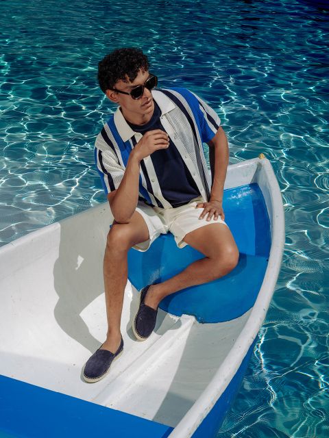 Ein Mann mit Sonnenbrille, gestreiftem Hemd und Shorts sitzt entspannt in einem kleinen weiß-blauen Boot auf dem klaren, glitzernden blauen Wasser.