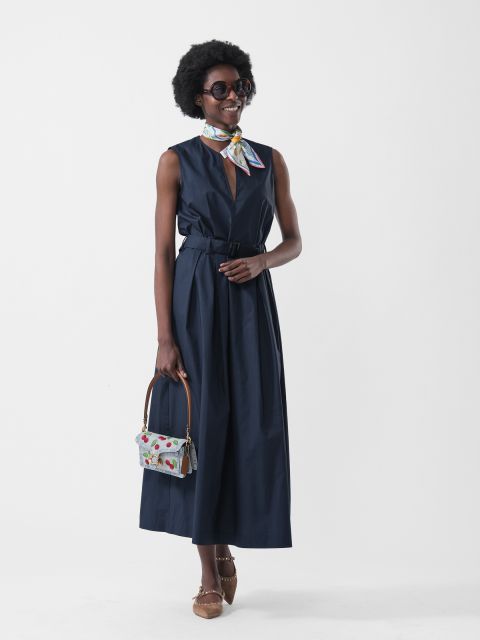Frau in einem ärmellosen marineblauen Kleid, einem bunten Schal, einer Sonnenbrille, einer gemusterten Handtasche und braunen Schuhen, vor einem weißen Hintergrund.