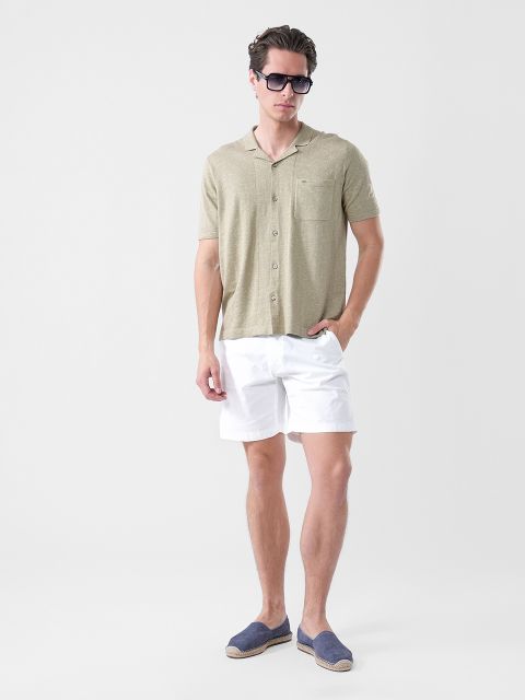 Ein Mann, der eine Sonnenbrille, ein hellolivfarbenes kurzärmeliges Hemd, weiße Shorts und marineblaue Espadrilles trägt, steht vor einem schlichten weißen Hintergrund.