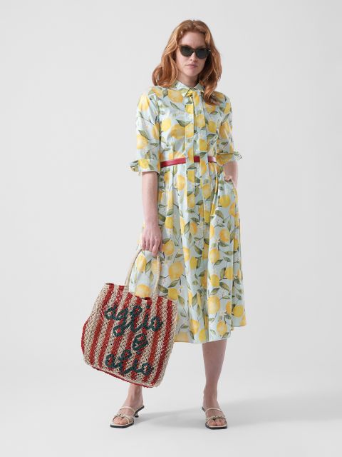 Frau in einem Kleid mit Zitronenmuster, Sonnenbrille und Sandalen hält eine rot-weiß gestreifte Tasche mit Schriftzug vor einem schlichten weißen Hintergrund.