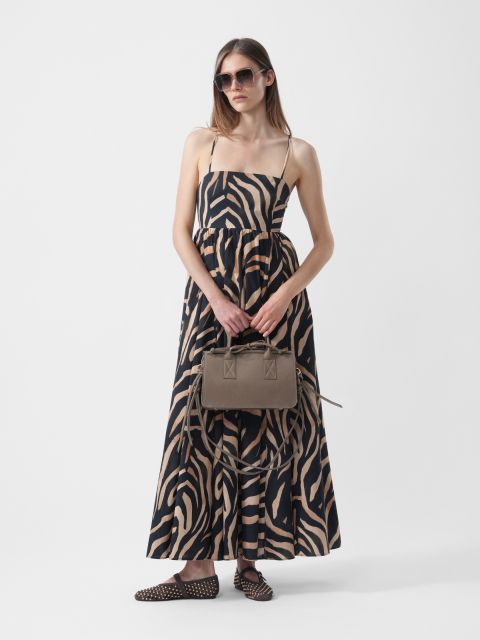 Frau trägt ein zebragestreiftes Maxikleid, Sonnenbrille und gepunktete Schuhe und hält eine taupefarbene Handtasche vor einem schlichten weißen Hintergrund.