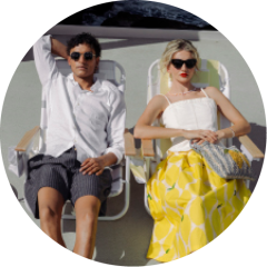 Ein Mann und eine Frau sitzen auf Liegestühlen in der Sonne. Der Mann trägt ein weißes Hemd und kurze Hosen, die Frau eine Sonnenbrille und einen gelb gemusterten Rock.