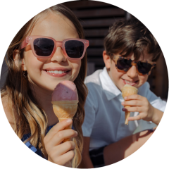 Zwei lächelnde Kinder mit Sonnenbrillen genießen an einem sonnigen Tag Eiswaffeln im Freien.