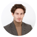 Junger Mann mit kurzen braunen Haaren, der einen grauen Blazer und einen hellbraunen Rollkragenpullover trägt und vor einem schlichten hellen Hintergrund posiert.