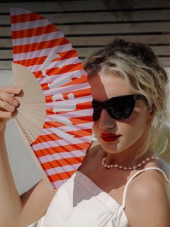 Eine Frau mit blonden Haaren und Sonnenbrille hält einen rot-weiß gestreiften Fächer in der Hand, trägt ein weißes Oberteil und eine Perlenkette und sitzt in der Sonne.