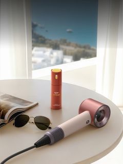 Auf einem runden Tisch neben einem Fenster, von dem aus man einen malerischen Blick auf die Küste hat, stehen ein Föhn, eine Sonnenbrille und eine kleine Sprühflasche.