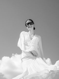 Eine Frau in einem wallenden weißen Kleid und einer großen Sonnenbrille lächelt unter einem wolkenverhangenen Himmel; das Foto ist in Schwarz-Weiß.