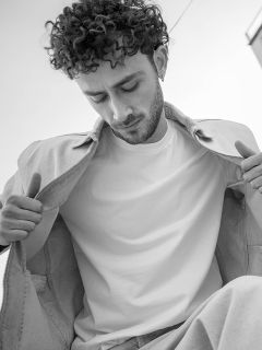 Schwarz-Weiß-Foto eines Mannes mit lockigem Haar, der nach unten schaut; er trägt eine leichte Jacke über einem schlichten weißen T-Shirt und hält die Jacke offen.