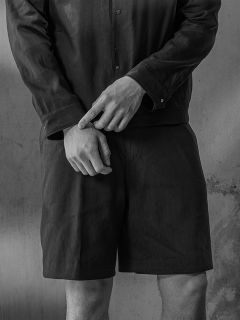 Schwarz-Weiß-Foto einer Person in einem Hemd und Shorts, die die Hände vor der Taille verschränkt hat und vor einer strukturierten Wand steht.