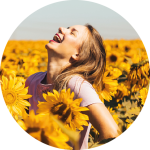 Eine Frau in einem rosa Hemd lacht fröhlich, während sie an einem sonnigen Tag in einem Feld voller blühender Sonnenblumen steht.