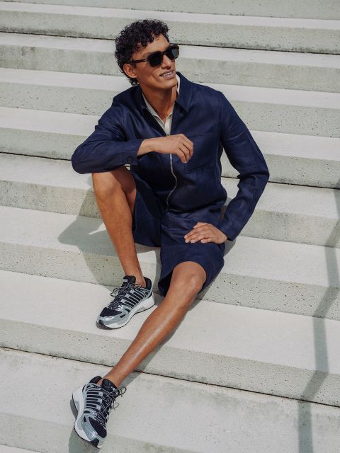 Ein Mann in marineblauen Shorts und Jacke, Sonnenbrille und Turnschuhen sitzt auf einer Betontreppe im Freien, lächelt und entspannt sich im hellen Sonnenlicht.