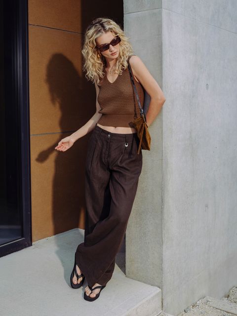 Eine Frau mit lockigem blondem Haar, die eine Sonnenbrille, ein braunes ärmelloses Oberteil, eine Hose mit weitem Bein und Sandalen trägt, steht an einer Betonwand im Sonnenlicht.