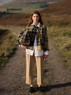 Eine Person steht auf einem Feldweg in einer hügeligen Landschaft und trägt eine karierte Jacke, ein weißes Hemd, eine beige Hose und schwarze Schuhe.