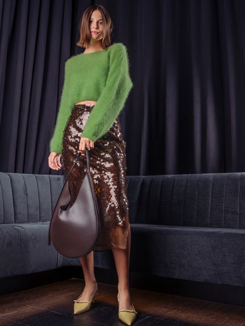 Eine Frau posiert in einem flauschigen grünen Pullover, einem braunen Midirock mit Pailletten und hellgelben Pumps und hält eine große, dunkelbraune Leder-Hobo-Tasche vor einem dunklen Vorhang in der Hand.