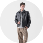 Ein Mann in einer schwarzen Lederjacke, einem gestreiften Hemd und einer beigen Hose steht mit einer Hand in der Tasche vor einem schlichten, hellen Hintergrund.